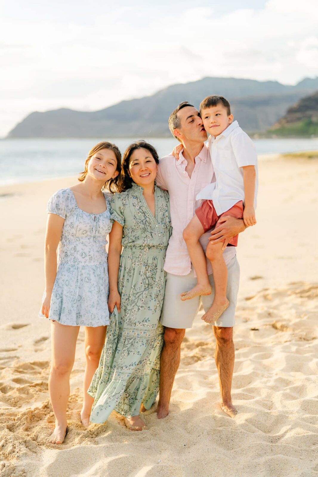 A smiling family of four stands barefoot on a sandy beach with mountains in the background. The father holds a young boy, while the mother and daughter stand close together, all wearing light, summery clothes.