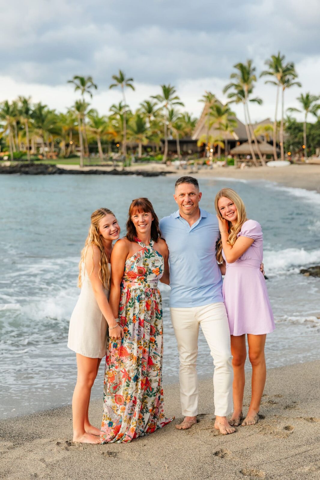 A smiling family of four stands barefoot on a sandy beach with palm trees and ocean waves in the background, wearing light summery clothes.