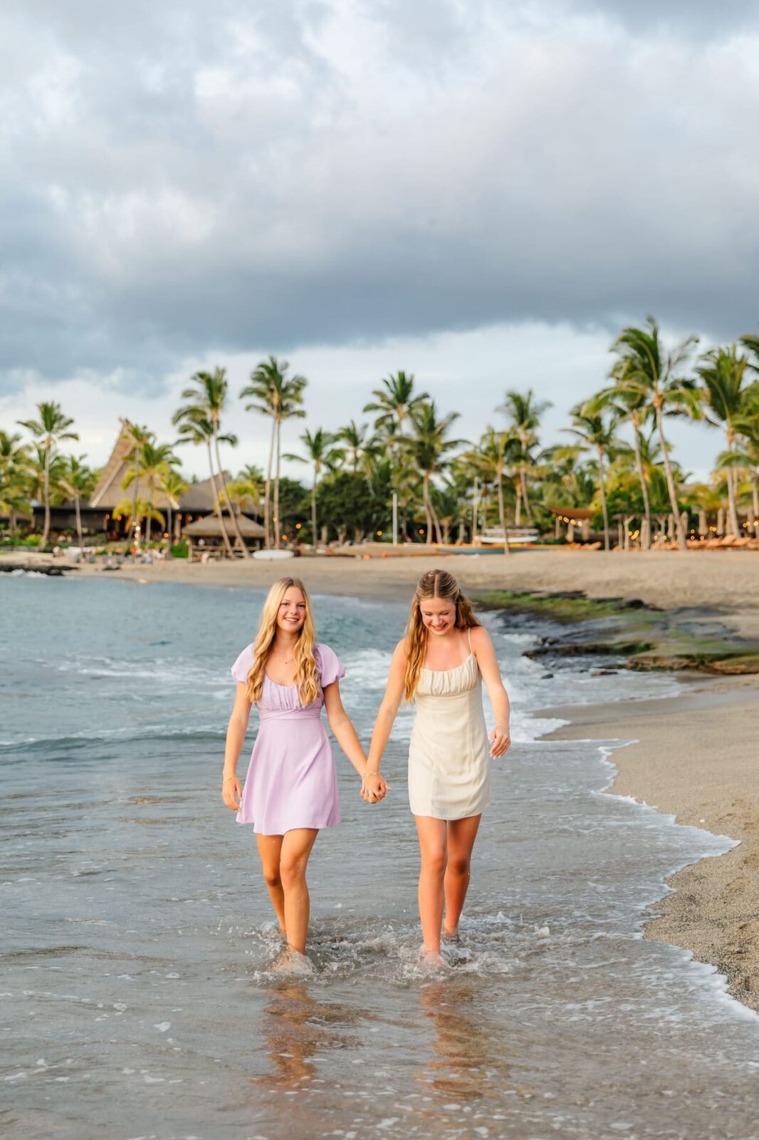 Two young women in light-colored dresses walk hand in hand along a tropical beach, smiling, with waves at their feet. Palm trees and a thatched-roof building are visible in the background under a cloudy sky.