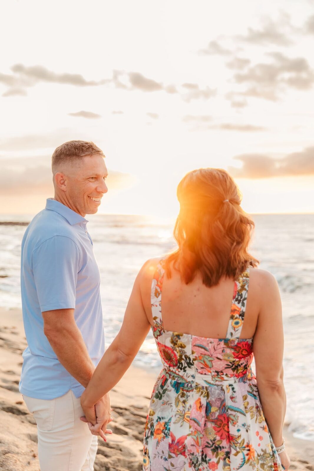 A couple holding hands and standing on a sandy beach at sunset. The man is smiling and wearing a light blue shirt, while the woman in a colorful floral dress faces the ocean, backlit by the sun.