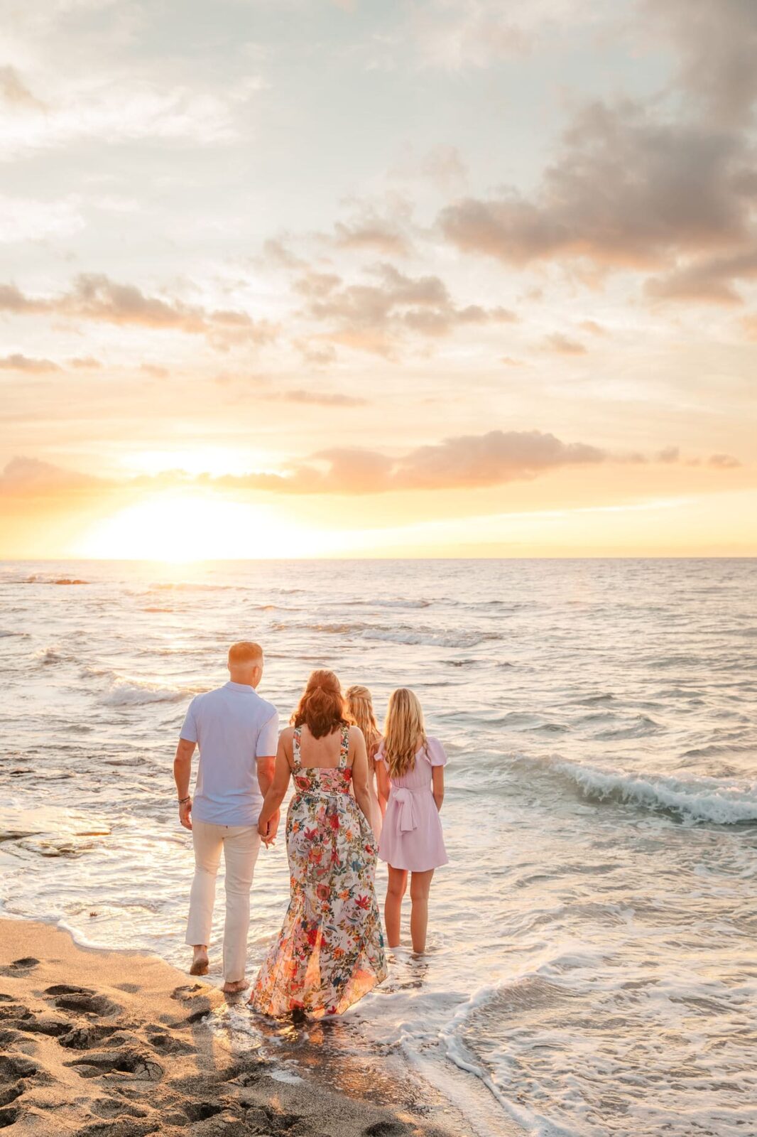 A family of four, seen from behind, stands at the edge of the ocean during sunset. The adults and two girls hold hands on the sandy beach, with waves gently touching their feet and the sky glowing orange and pink.