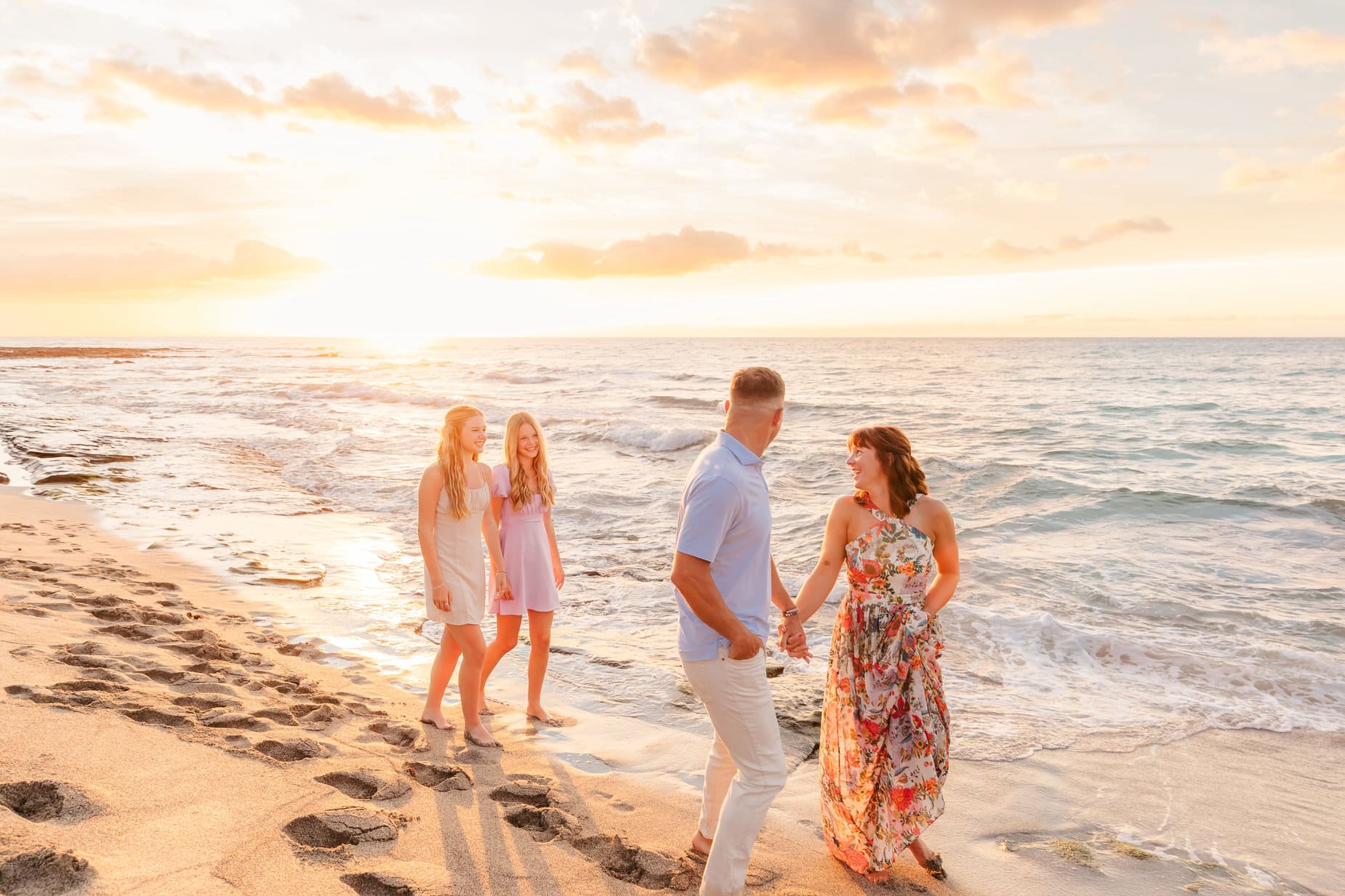 A family of four walks along the beach at sunset. The parents hold hands and smile at each other, while two teenage girls walk ahead, laughing. Gentle waves wash onto the sandy shore under a colorful sky.
