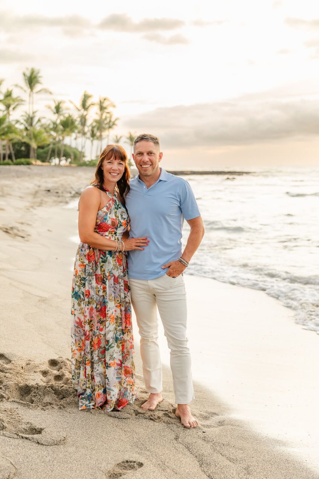A woman in a floral dress and a man in a light blue shirt and white pants smile while standing barefoot on a sandy beach with palm trees and waves in the background at sunset.