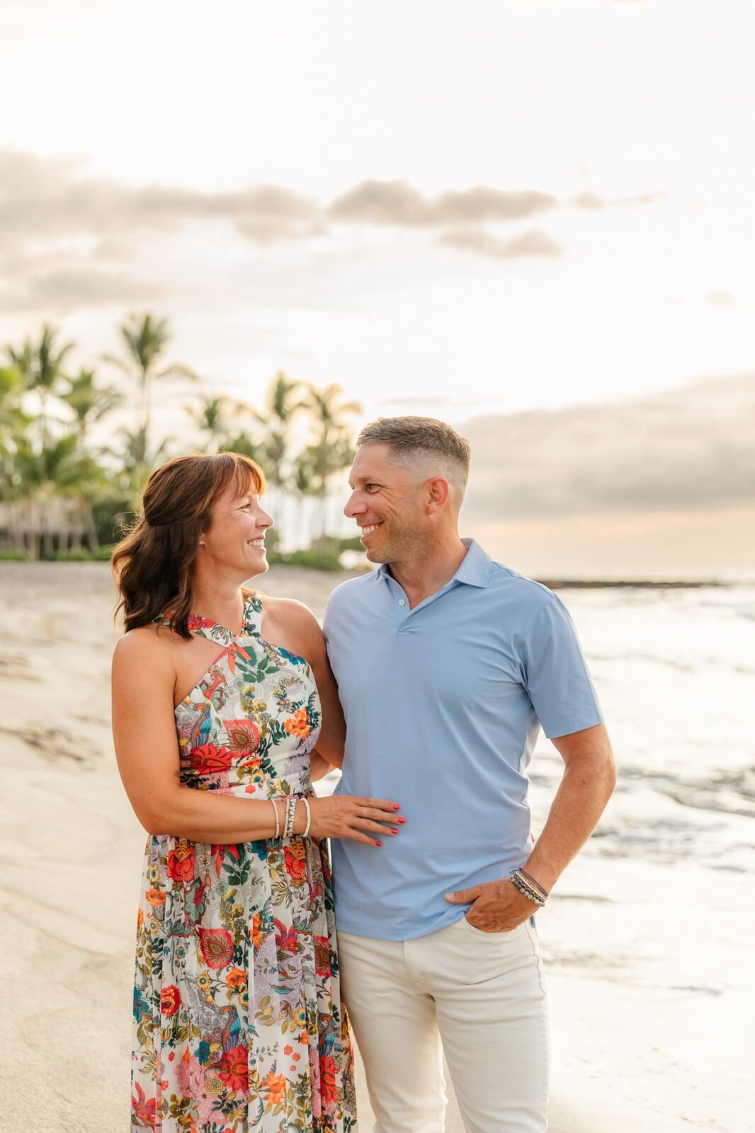A smiling couple stands close together on a sandy beach at sunset. The woman wears a colorful floral dress, and the man wears a light blue polo shirt and white pants. Palm trees and gentle waves are in the background.