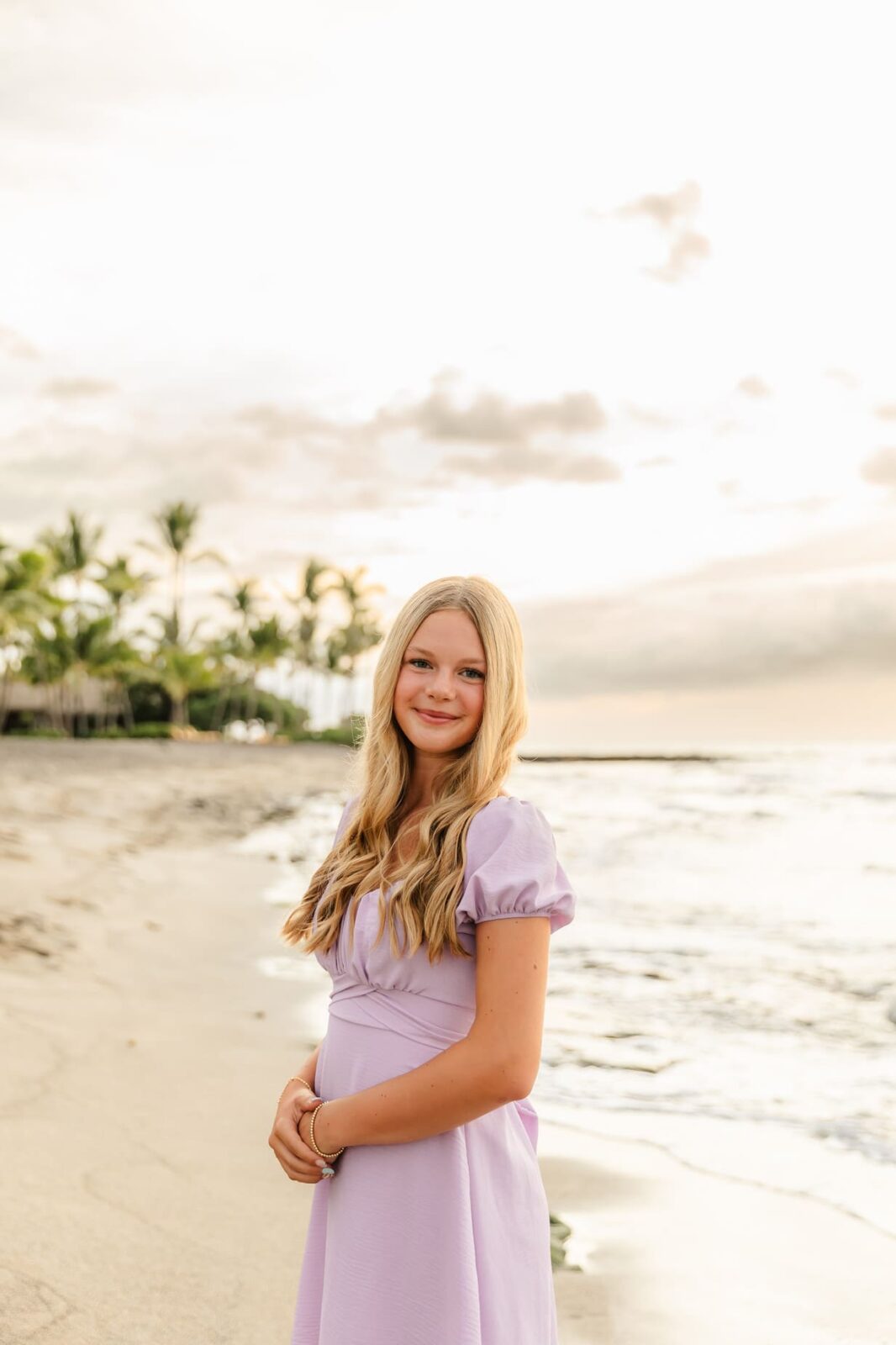 A young girl with long blonde hair in a light purple dress stands on a sandy beach near the ocean, smiling softly. Palm trees and a pastel sunset sky are visible in the background.
