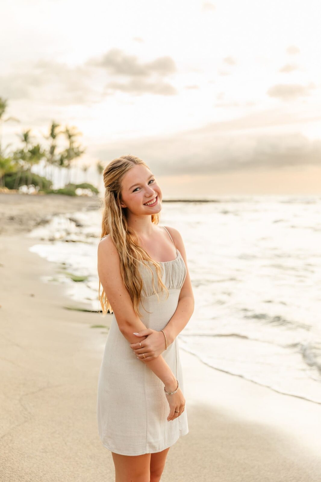 A young woman with long blonde hair, wearing a light beige sundress, smiles while standing on a sandy beach at sunset with gentle waves and palm trees in the background.