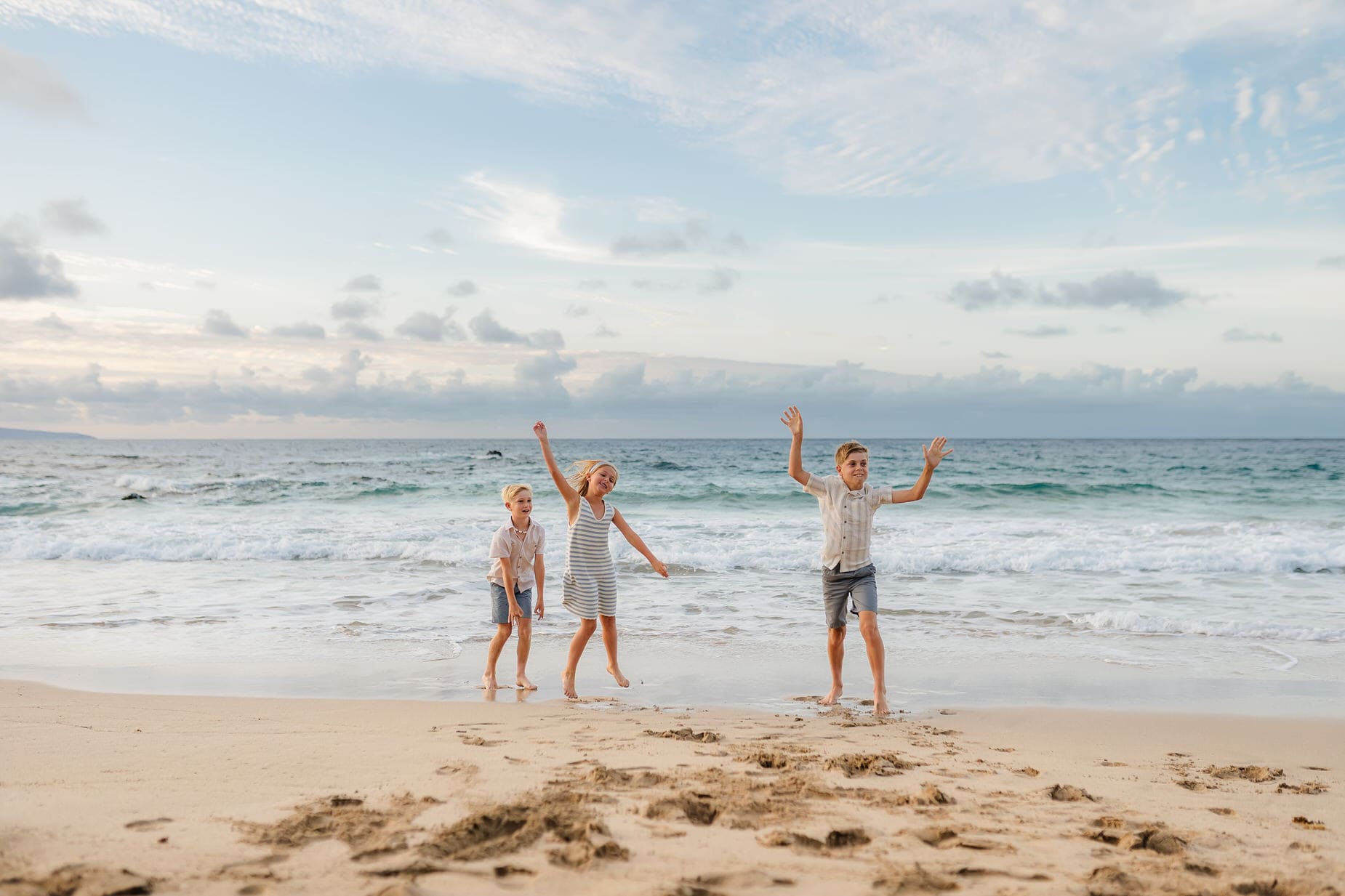 Three children stand and play on a sandy Maui beach near the shoreline, enjoying gentle waves beneath a cloudy sky—capturing the joy and magic perfect for professional family portraits in Maui.