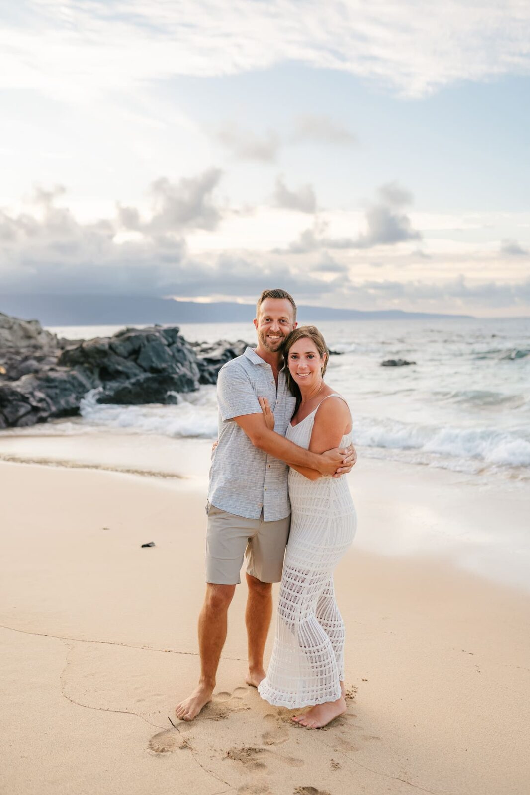 A couple stands barefoot on a sandy beach, smiling and embracing near the shoreline—perfect inspiration for professional family portraits Maui, with gentle waves, rocks, and a cloudy sky in the background.