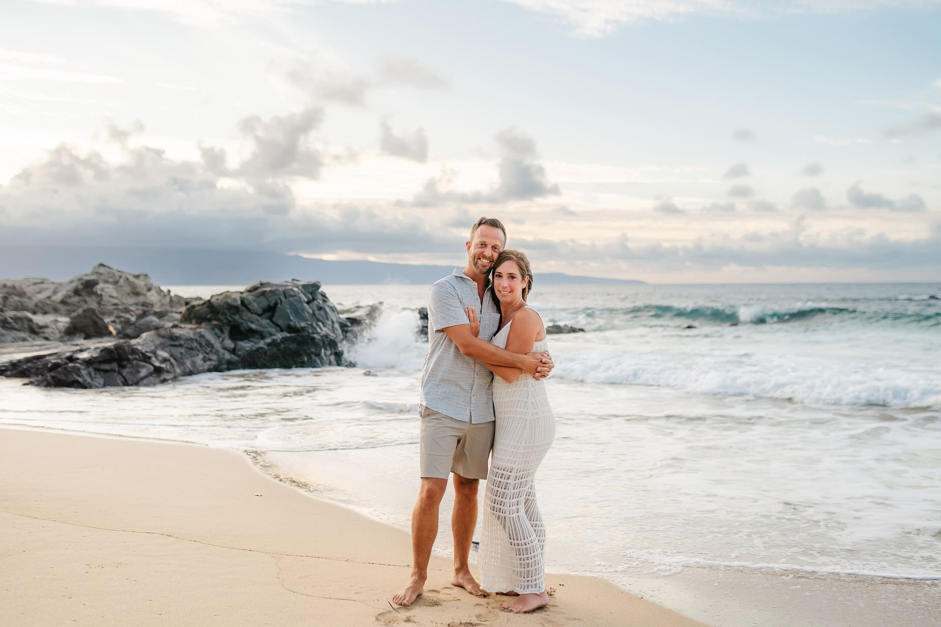 A couple stands barefoot on a sandy beach, embracing and smiling at the camera with waves crashing behind them and rocky cliffs to the left under a cloudy sky at sunset—perfect for professional family portraits in Maui.