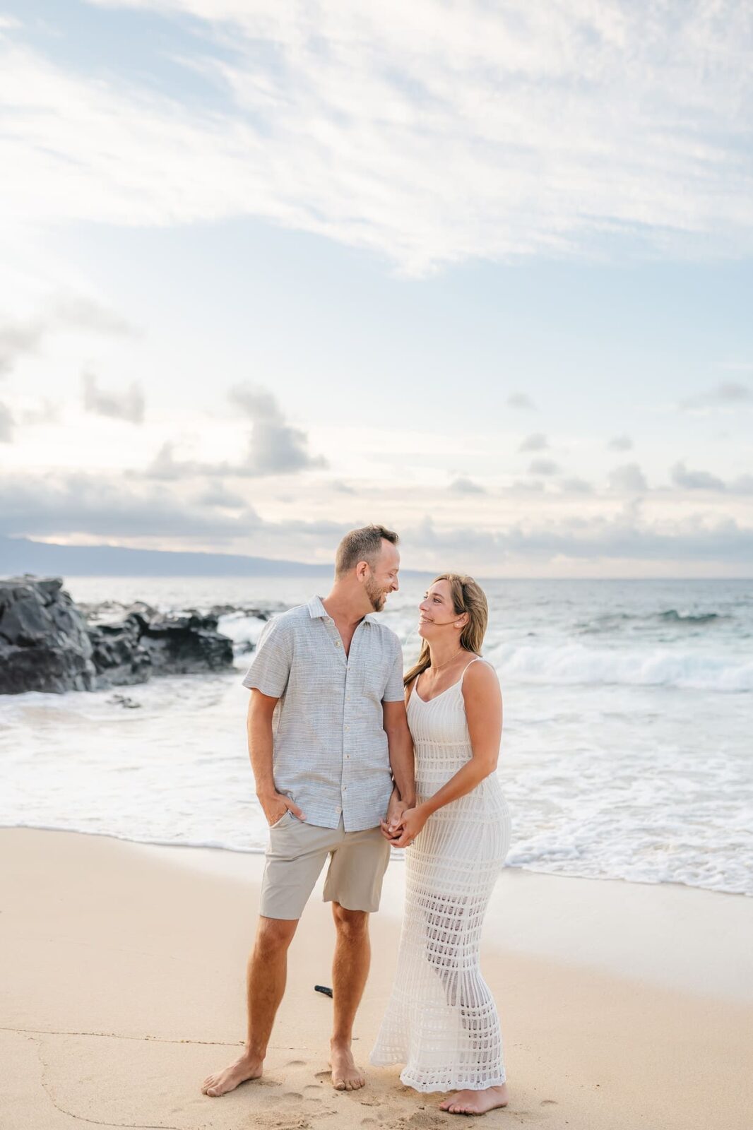 A man and woman stand barefoot on a sandy beach, smiling at each other while holding hands. Waves crash behind them under a partly cloudy sky, capturing the serene beauty found in professional family portraits Maui is known for.