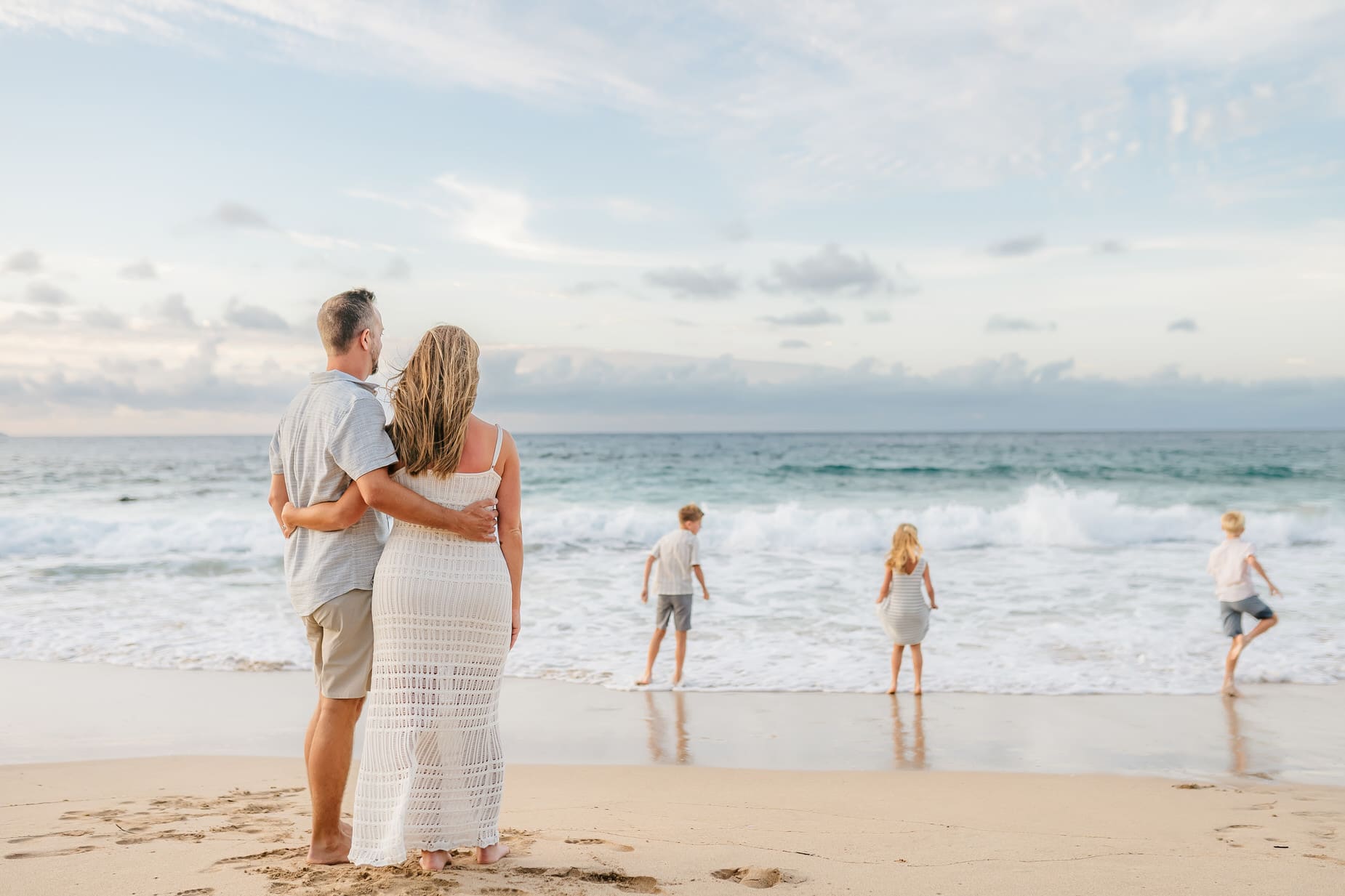 A couple stands arm in arm on a sandy beach, watching three children play near the shoreline as waves roll in under a partly cloudy sky—capturing the essence of professional family portraits Maui style.