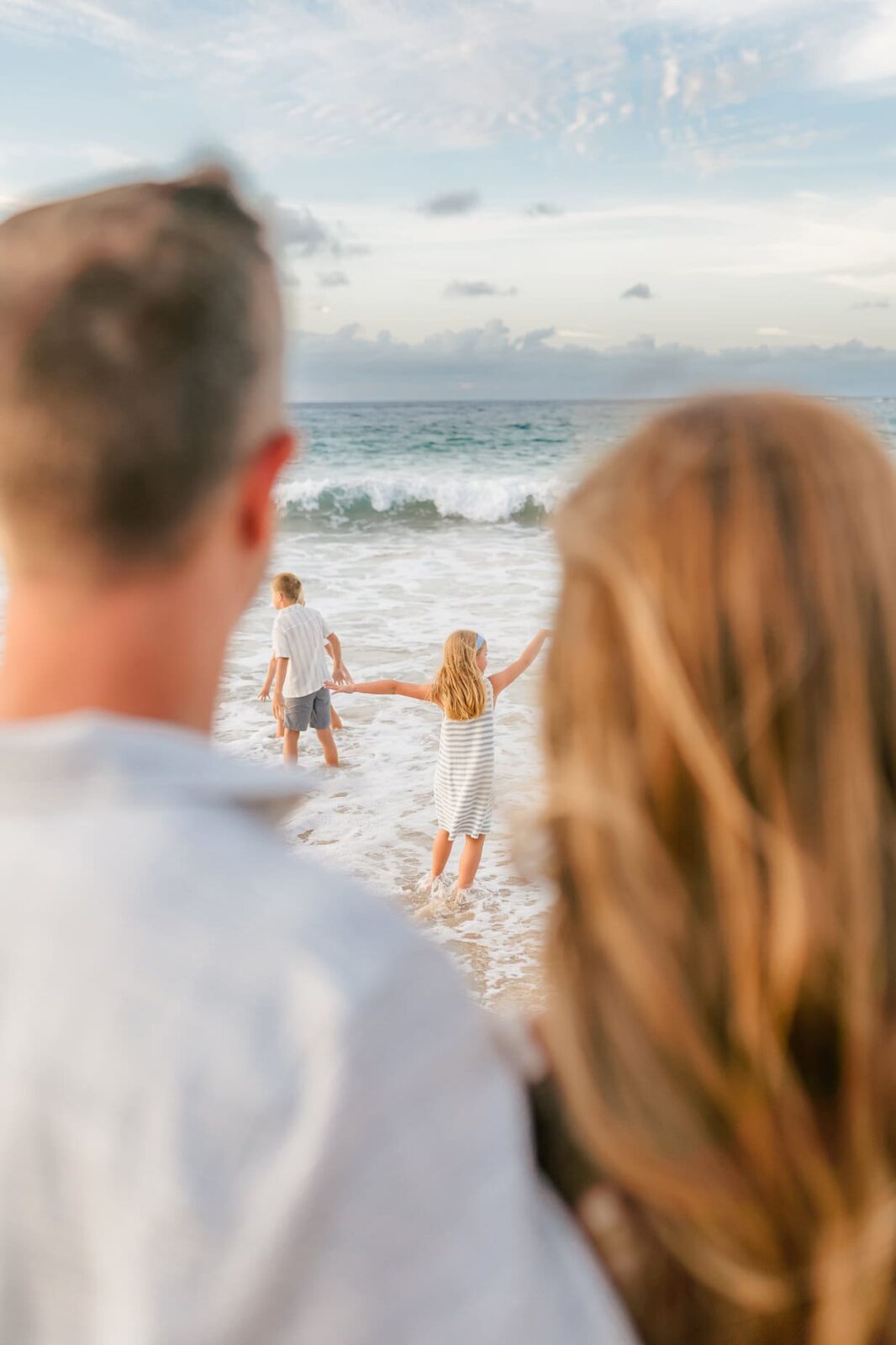 A young girl and boy play at the edge of the ocean, waves rolling in, while two adults watch from behind. The scene is bright and joyful, a perfect moment for professional family portraits in Maui under a cloudy blue sky.