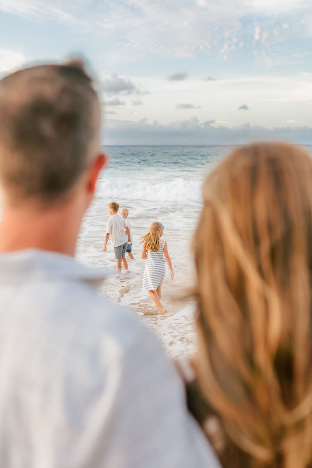 Two adults stand in the foreground, watching two children walk toward the ocean on a sandy beach under a partly cloudy sky—perfect for professional family portraits in Maui, capturing cherished moments of togetherness by the sea.
