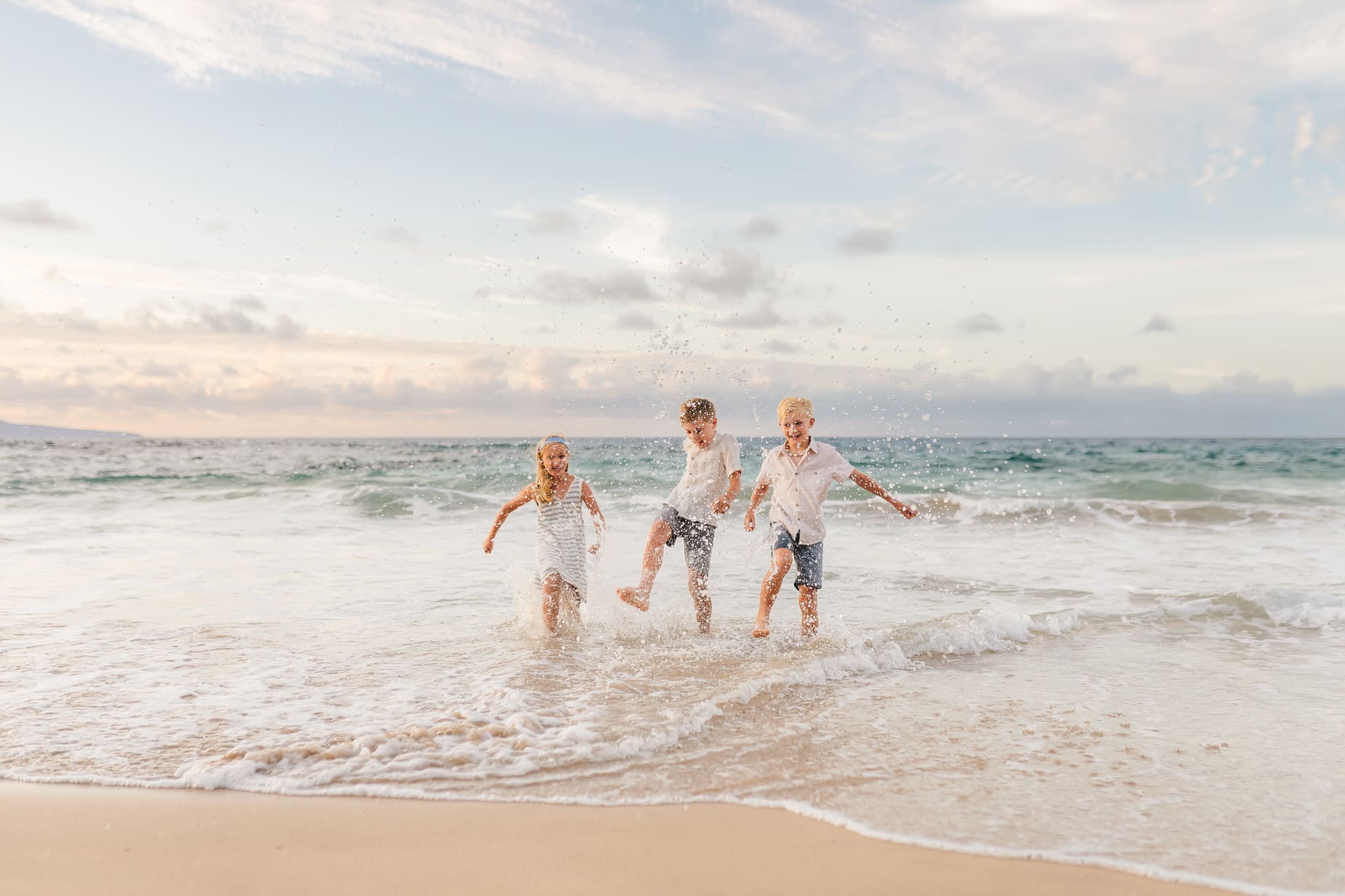 Three children play and splash in shallow ocean waves at the beach during sunset, laughing and kicking water—a candid moment perfect for professional family portraits Maui with the vibrant sky and sea as a stunning backdrop.