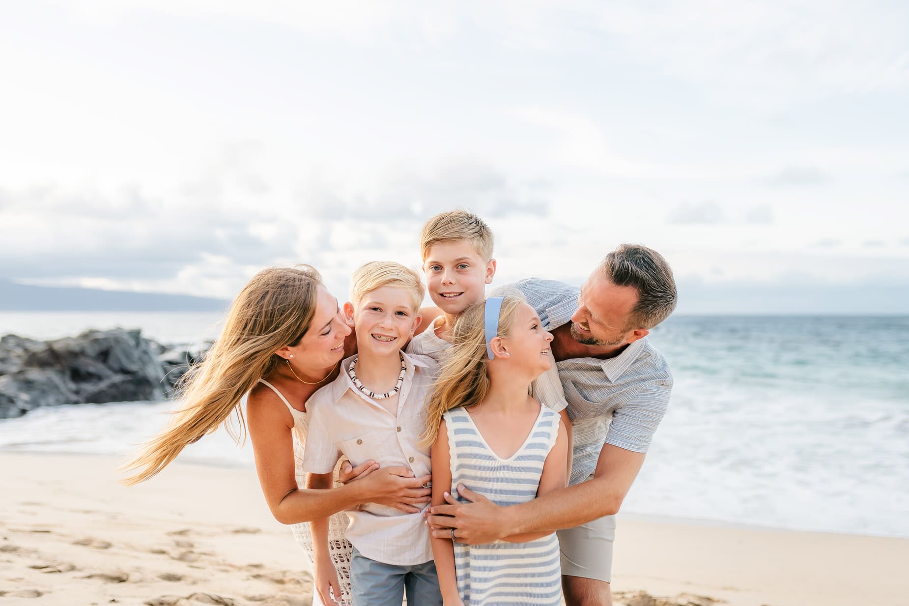 A family of five, including two adults and three children, smiles and embraces on a sandy beach with the ocean and rocks in the background. Capture precious memories with professional family portraits Maui under a partly cloudy sky.