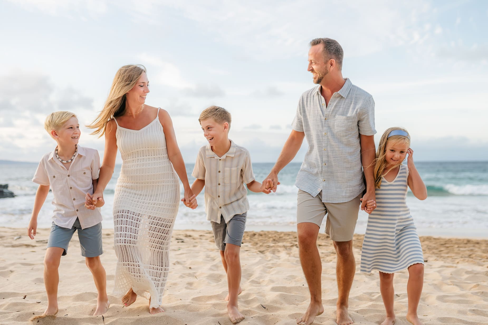 A family of five walks hand-in-hand on a sandy beach in Maui, smiling and enjoying the day. The parents and three children wear light-colored summer clothes; the ocean and sky frame this perfect moment for professional family portraits Maui.