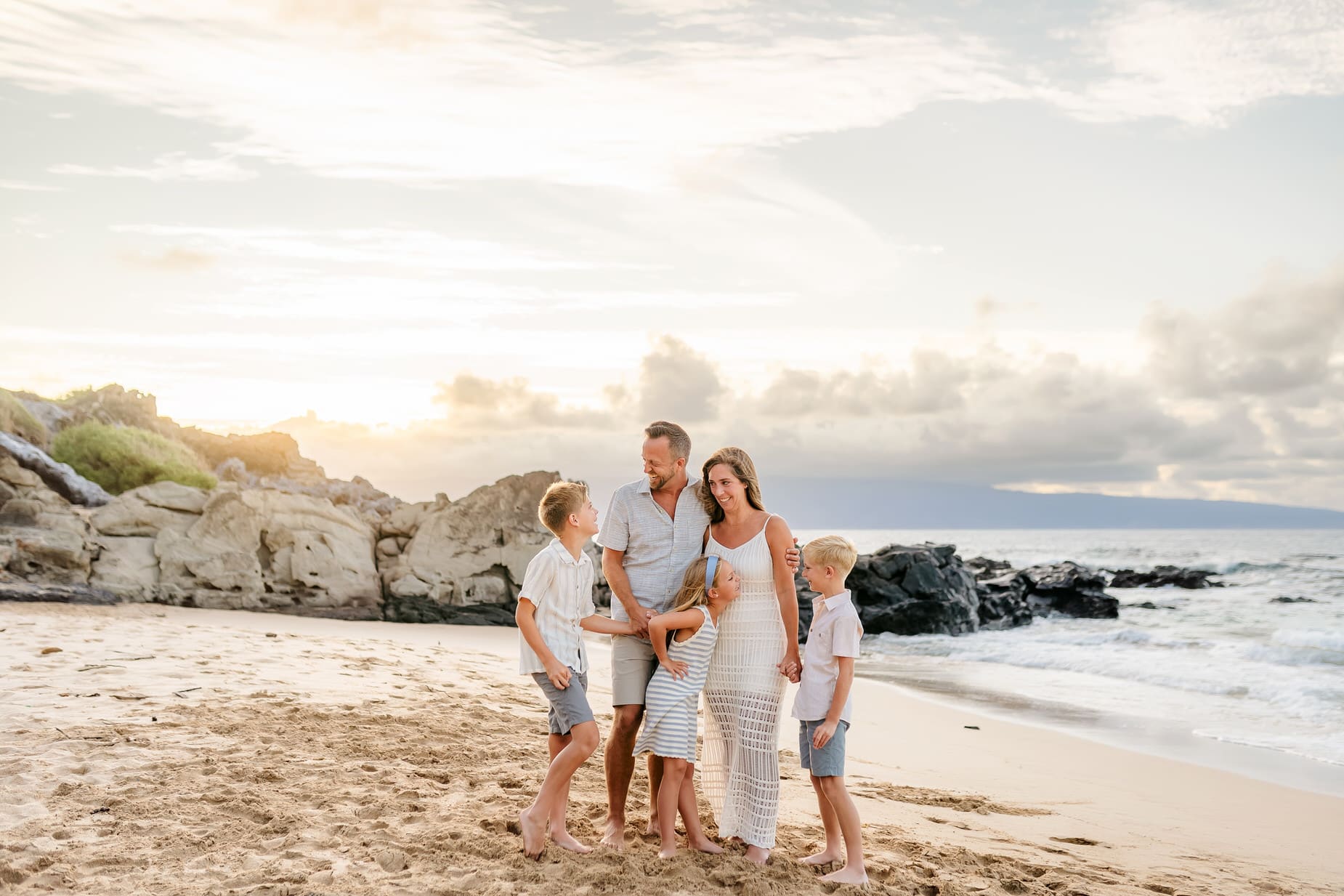 A family of five stands barefoot on a sandy beach at sunset, smiling and interacting with each other. Rocky cliffs and gentle waves are in the background under a partly cloudy sky, capturing the essence of professional family portraits Maui offers.