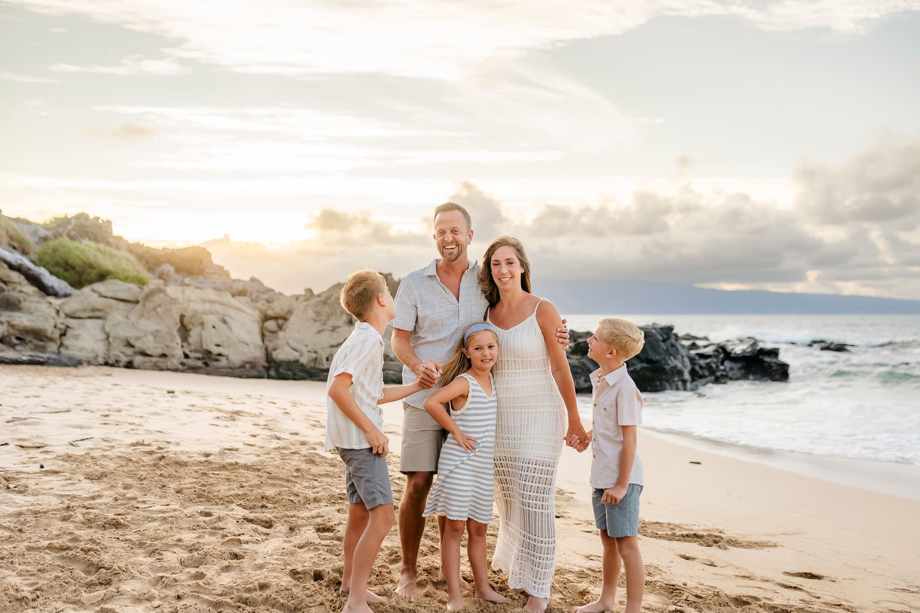 A smiling family of five stands barefoot on a sandy beach near rocky cliffs at sunset. Capturing professional family portraits in Maui, the parents are in the center with their three young children, all dressed in light summer clothes, enjoying their time together.
