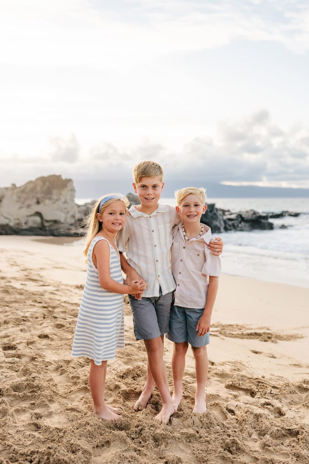 Three children, two boys and a girl, stand barefoot on a sandy beach near the ocean. Smiling together in front of rocks, they capture a joyful moment—perfect for professional family portraits in Maui.
