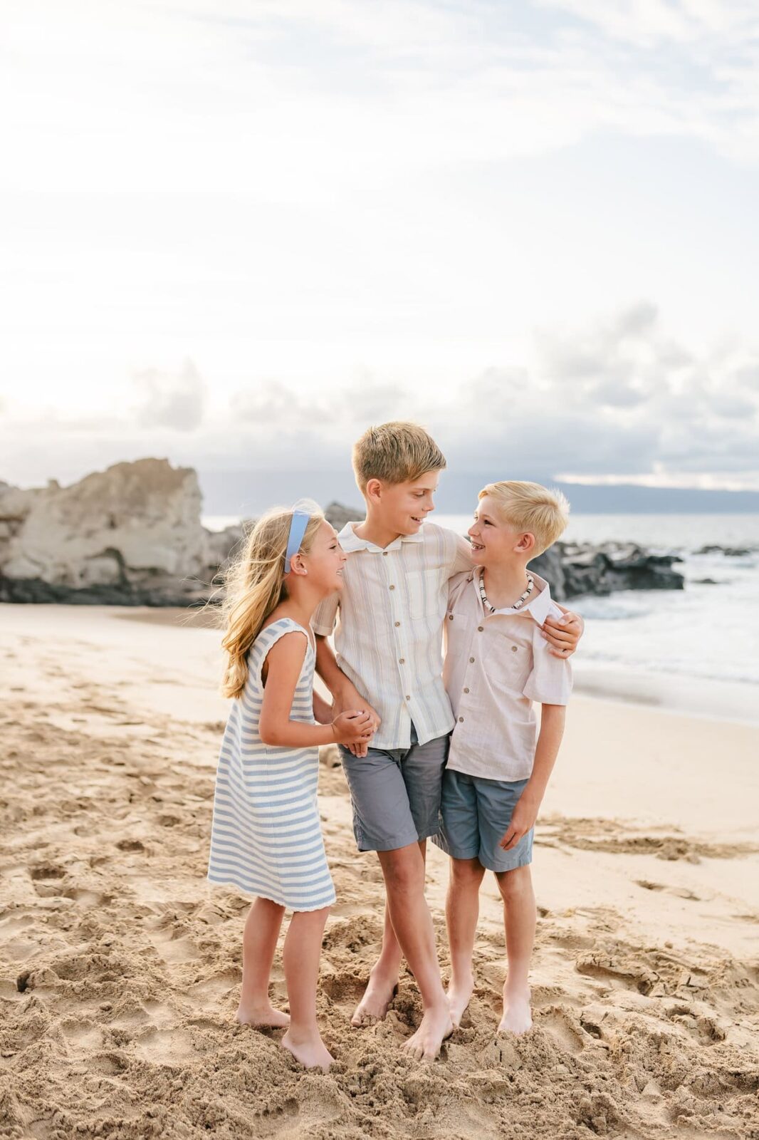 Three children stand barefoot on a sandy beach, smiling and laughing together. Capturing the joy of childhood, this moment is perfect for professional family portraits in Maui, with rocky outcrops and the ocean as a scenic backdrop.
