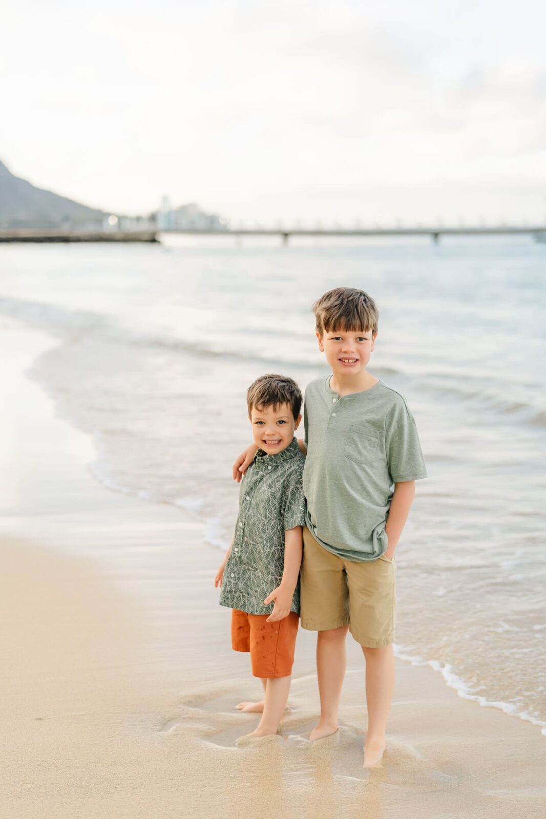 Two young boys stand barefoot on a sandy beach near the water’s edge, smiling at the camera during sunrise family photos on Oahu. One wears an olive green shirt and tan shorts, the other a patterned green shirt and orange shorts. A pier and mountain are behind them.