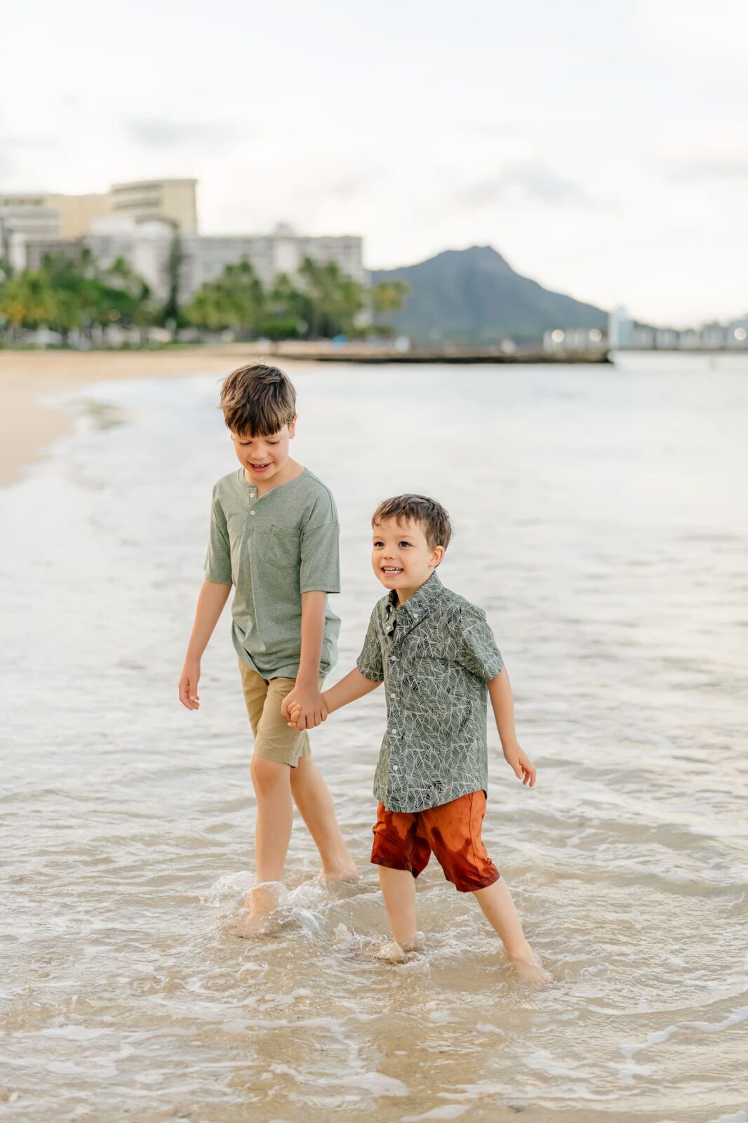 Two young boys walk hand in hand along the shoreline at sunrise, smiling as they wade through shallow water. Perfect for family photos on Oahu, with buildings, palm trees, and a mountain visible in the background.