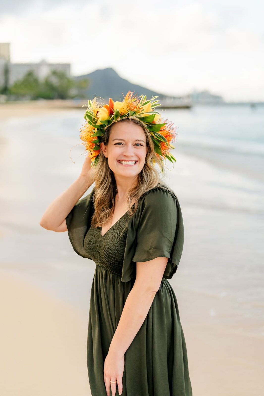 Smiling woman in a dark green dress and bright flower crown stands on a sandy beach with gentle waves, touching her crown—perfect for sunrise family photos on Oahu. A mountain and buildings are visible in the distant background.