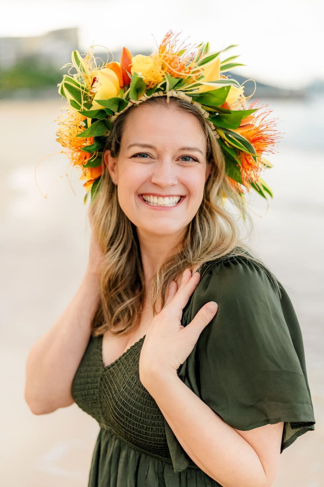 A smiling woman with long blond hair wears a green dress and a vibrant flower crown, standing on a beach at sunrise—perfect for capturing family photos on Oahu with the blurred shoreline in the background.