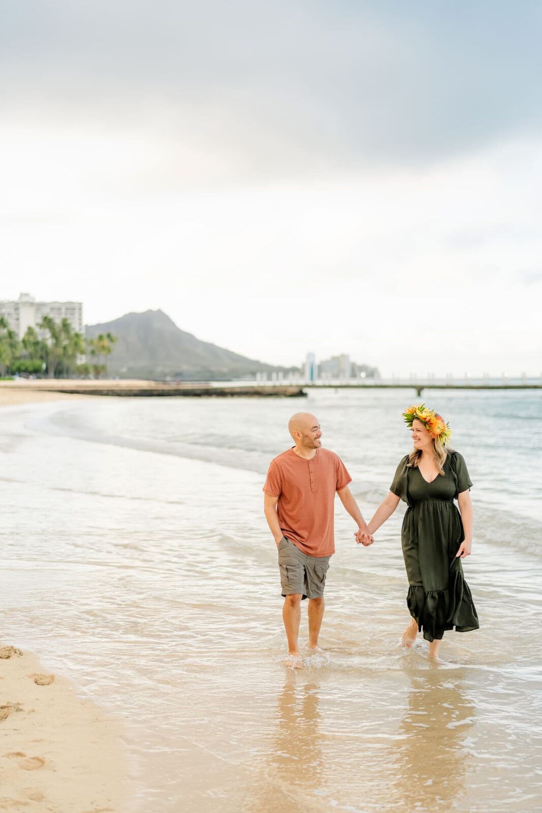 A couple holds hands while walking along a sandy beach with gentle waves at sunrise—perfect for family photos on Oahu. The man wears a rust t-shirt and shorts; the woman wears a dark dress and flower crown. Palm trees, buildings, and a mountain are in the background.
