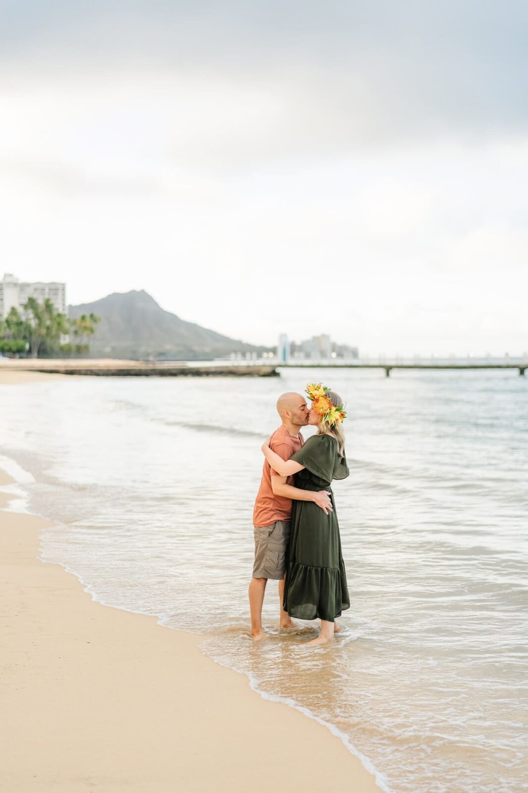 A couple stands barefoot on a sandy beach, embracing and kissing near the shoreline at sunrise. The woman wears a green dress and a sunflower crown—perfect inspiration for sunrise family photos on Oahu, with city buildings, mountains, and a pier in the background.