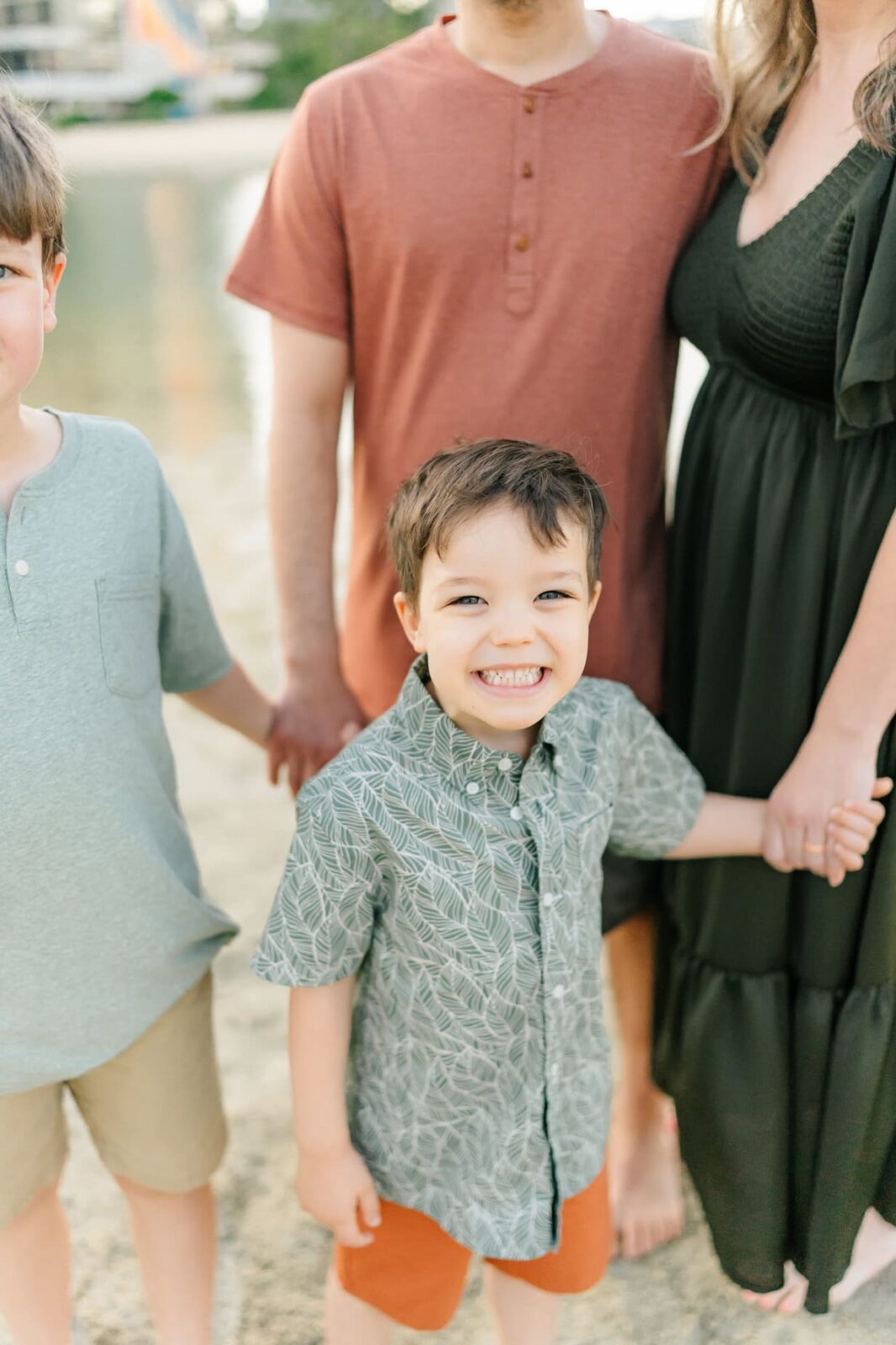 A young boy smiles at the camera while holding hands with two adults and another child at the beach, capturing a joyful moment during sunrise family photos on Oahu. The group stands on sand near water, with only the adults’ lower faces and bodies visible.
