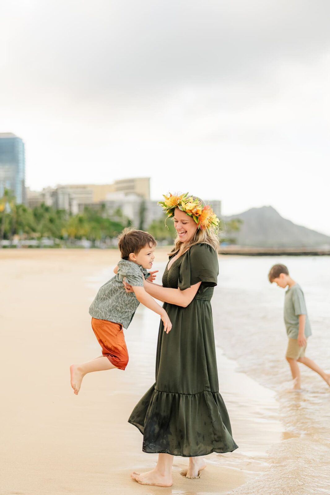 A woman in a dark green dress and flower crown smiles while holding a laughing child on a sandy beach during sunrise family photos on Oahu. Another child walks at the water’s edge, with buildings and a mountain in the background.