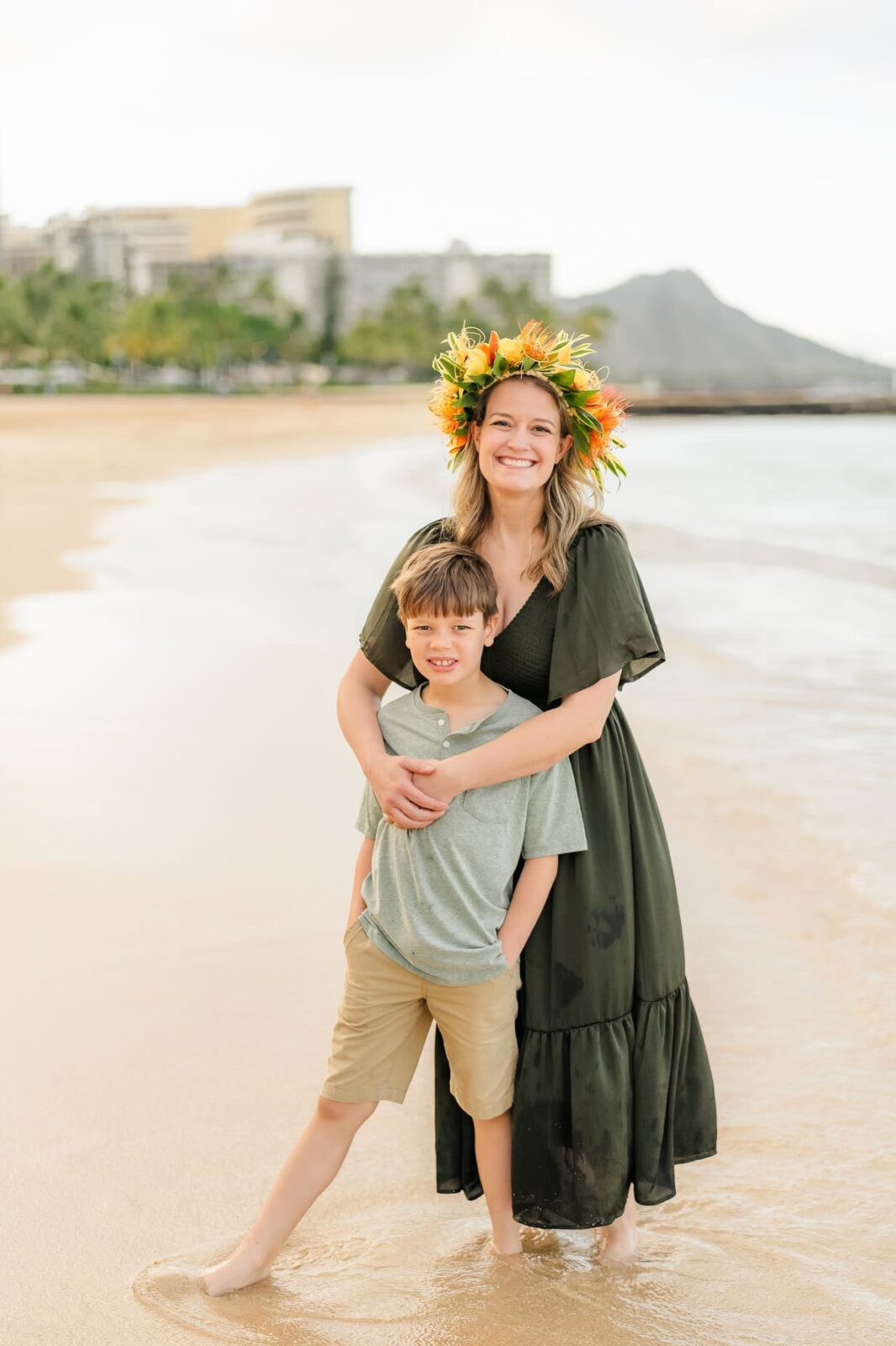 A woman in a flower crown and dark dress stands on a sandy beach with her arms around a smiling boy in green, capturing sunrise family photos on Oahu with ocean waves and distant buildings in the background.