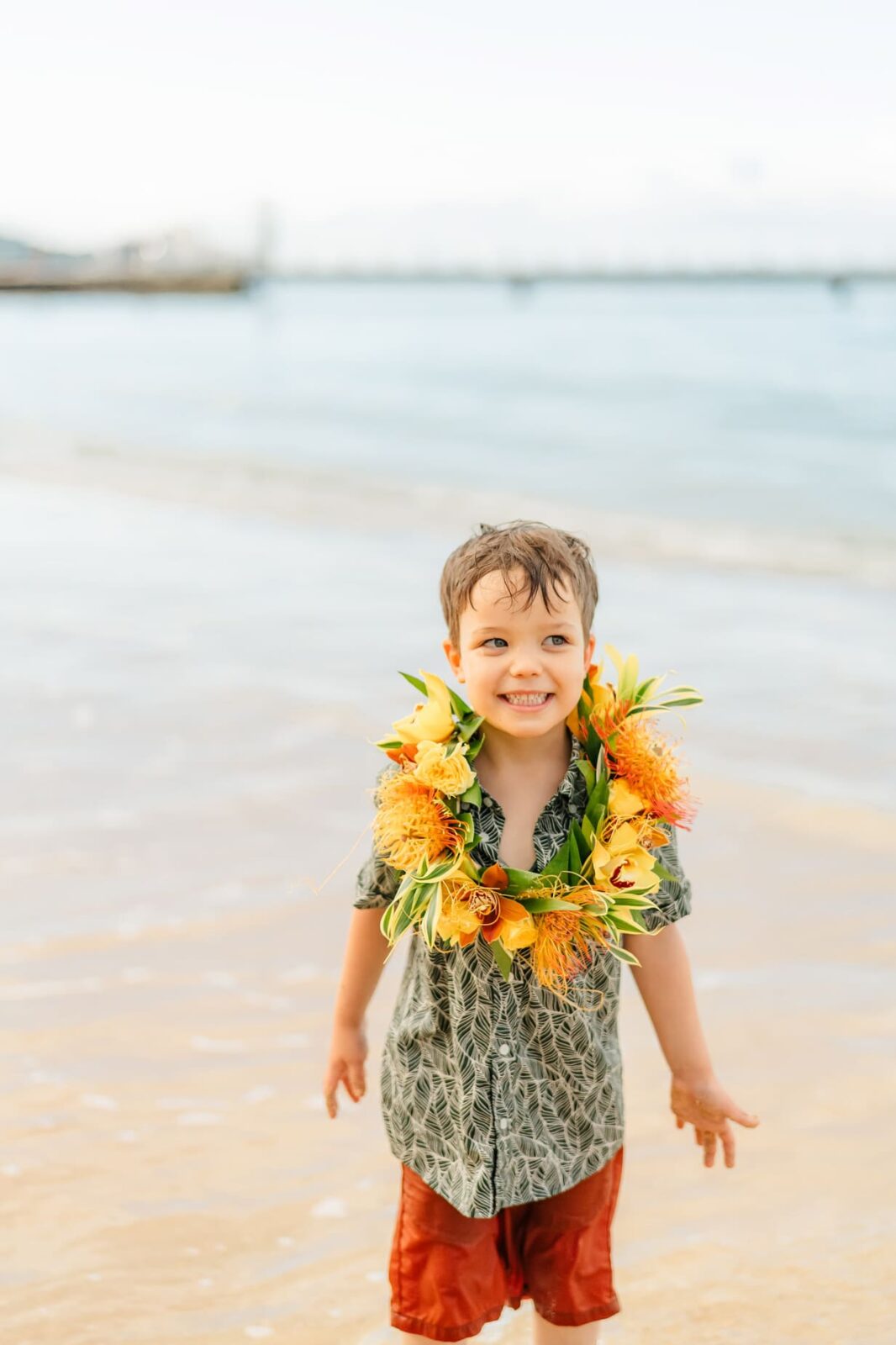 Smiling young boy wearing a floral lei, wet hair, patterned shirt, and red shorts stands on the sandy shore with ocean waves in the background, perfect for sunrise family photos on Oahu.