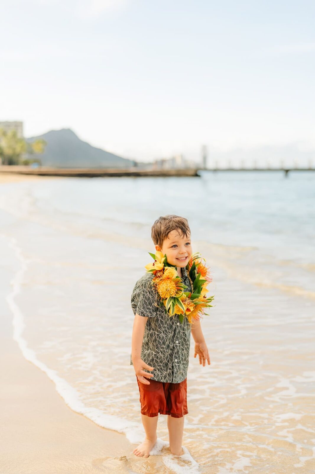 A young boy wearing a floral lei, patterned shirt, and red shorts smiles while standing barefoot on a sandy beach at sunrise on Oahu, with gentle waves, palm trees, and a distant mountain in the background—perfect for family photos.