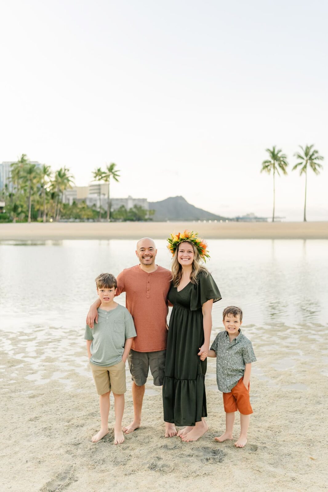 A smiling family of four stands barefoot on a sandy beach with palm trees, water, and a distant mountain in the background, enjoying sunrise family photos on Oahu. The mother wears a flower crown; the father and two boys wear casual summer clothes.