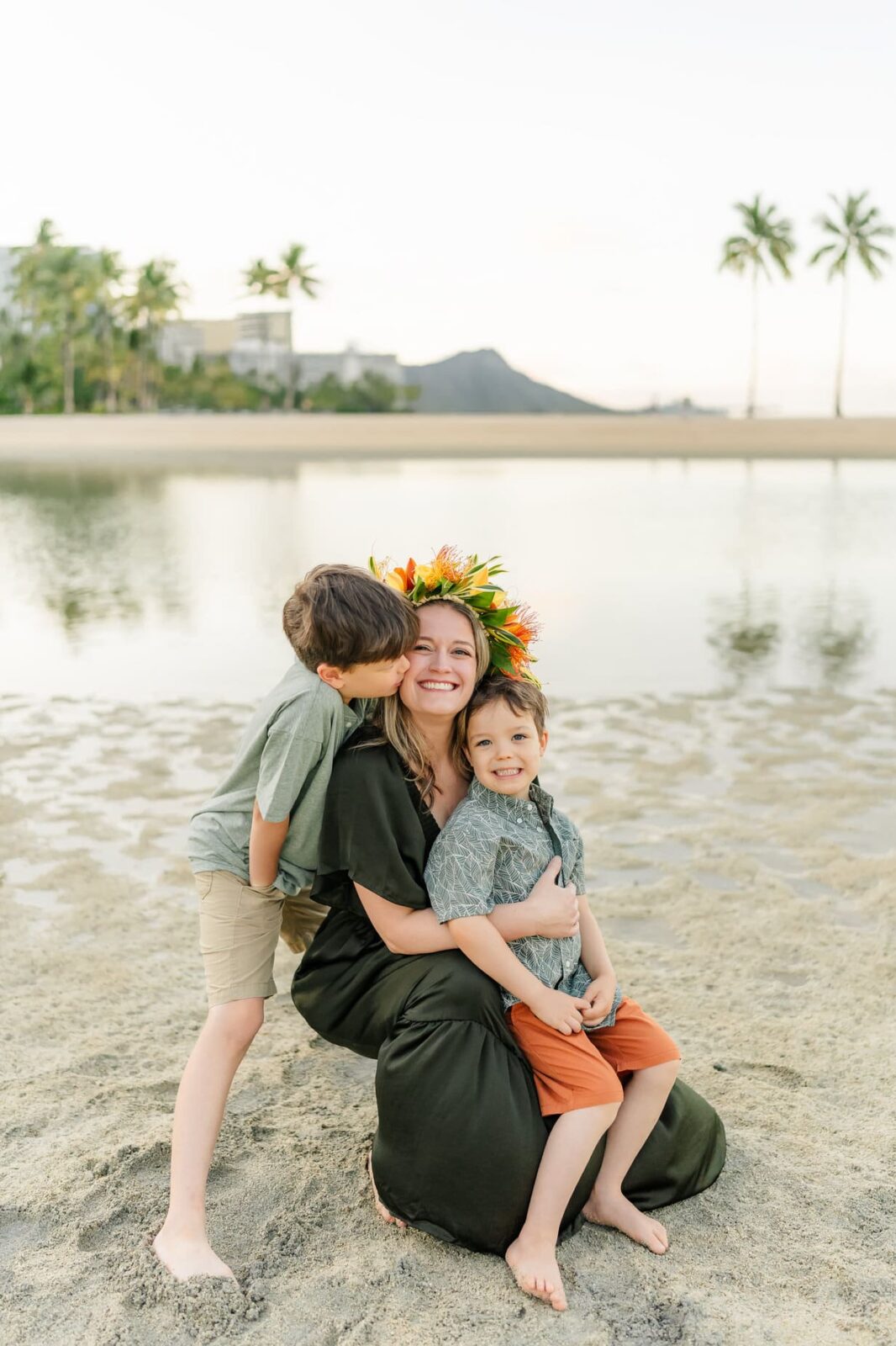 A woman wearing a flower crown hugs two young boys on a sandy beach during sunrise family photos on Oahu. One boy kisses her cheek while the other smiles at the camera, with palm trees, water, and a mountain in the background.