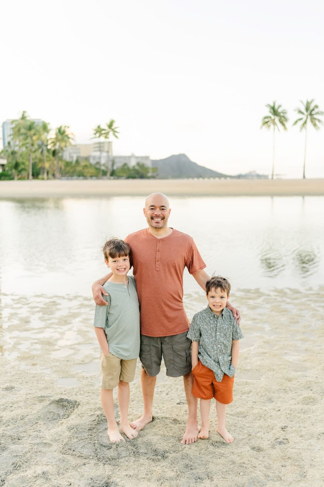 A man stands on a sandy beach with his arms around two young boys during sunrise family photos on Oahu. They are barefoot, smiling in casual clothes, with palm trees and a mountain visible across the water.