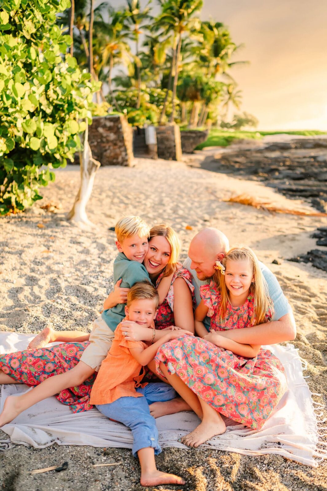 A family of five sits on a blanket at the beach, smiling and hugging each other. They are surrounded by palm trees and greenery, with golden sunlight illuminating the scene.