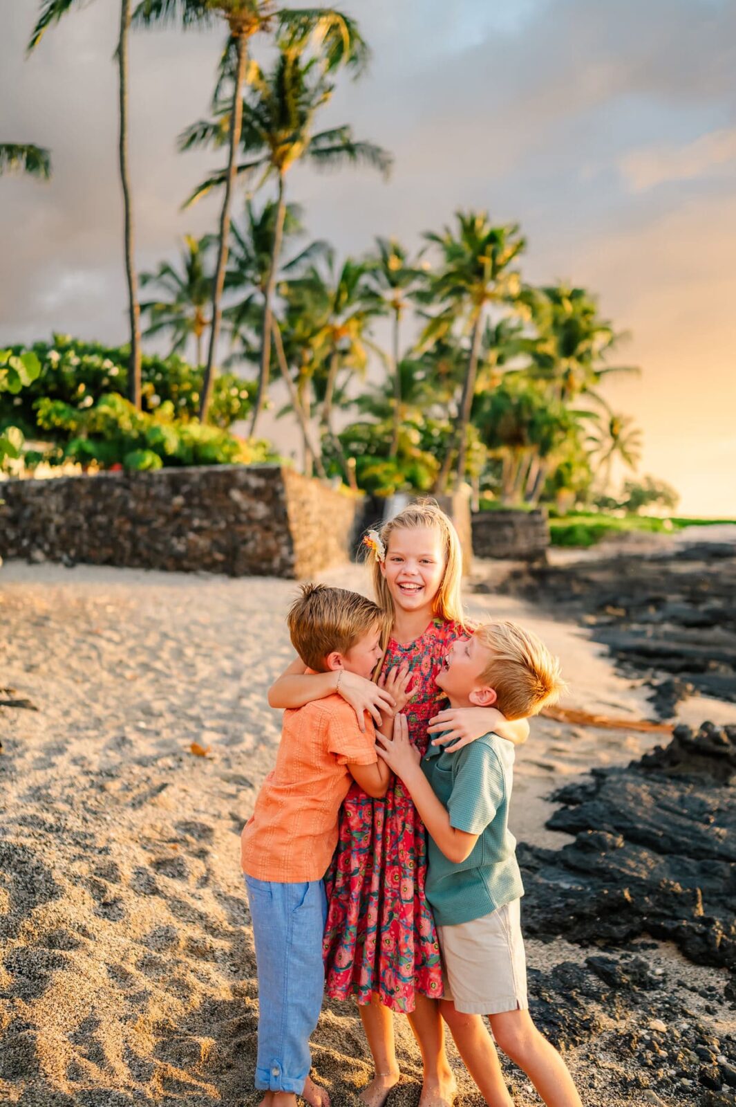 Three young children laugh and hug each other on a sandy beach at sunset, surrounded by palm trees and lush greenery. The girl in the middle wears a floral dress, while the two boys wear shorts and shirts.