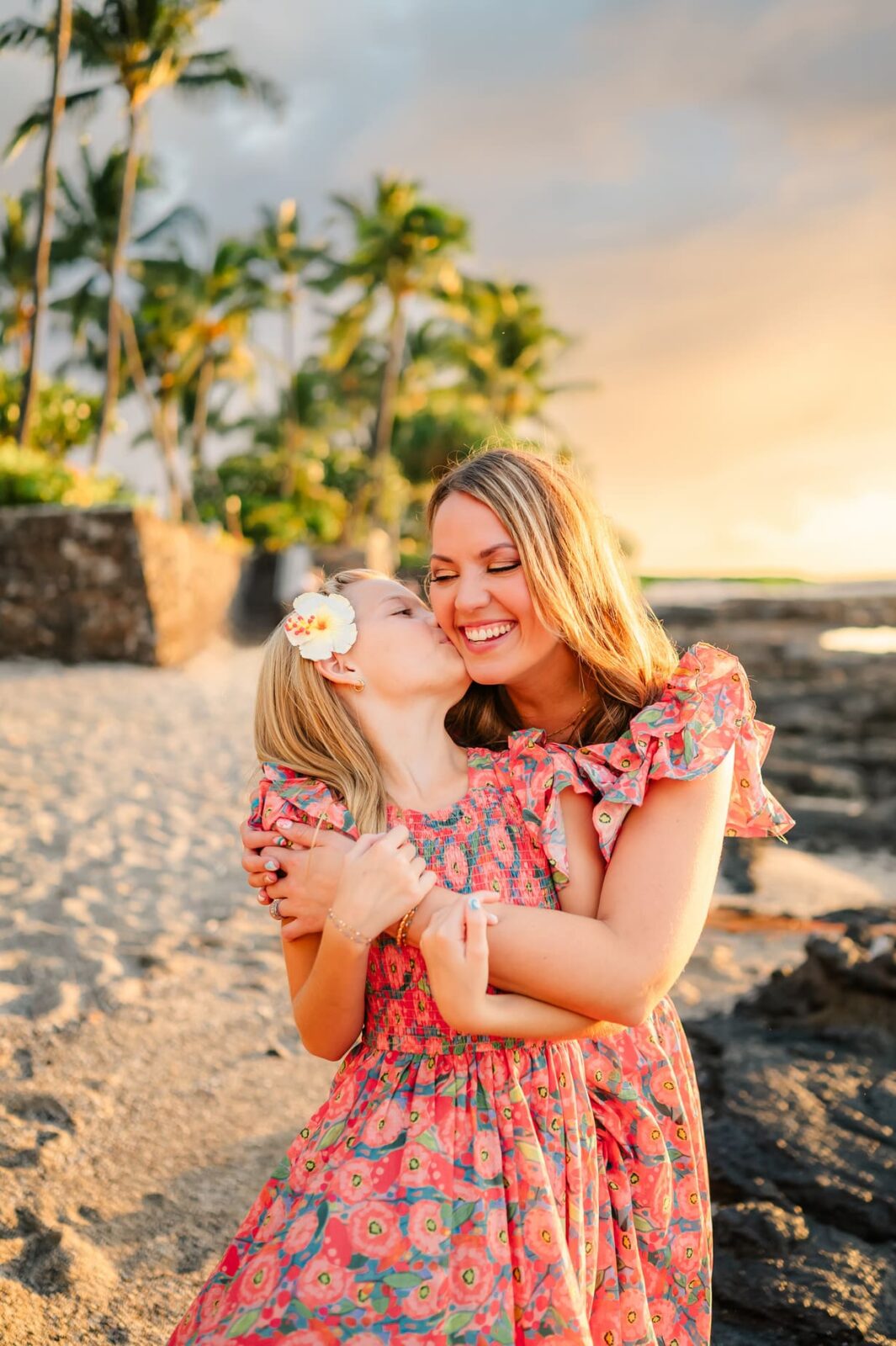 A woman and a young girl wearing matching floral dresses smile and hug on a tropical beach at sunset. The girl is kissing the woman’s cheek. Palm trees and stone walls appear in the background.