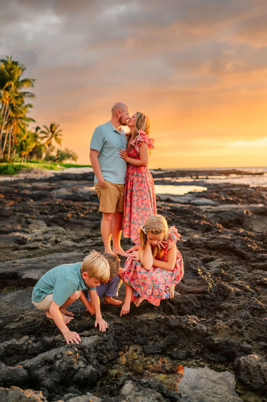 A couple kisses while standing on black rocks at sunset as two young children play and look at tide pools nearby. Palm trees and a colorful sky are in the background.