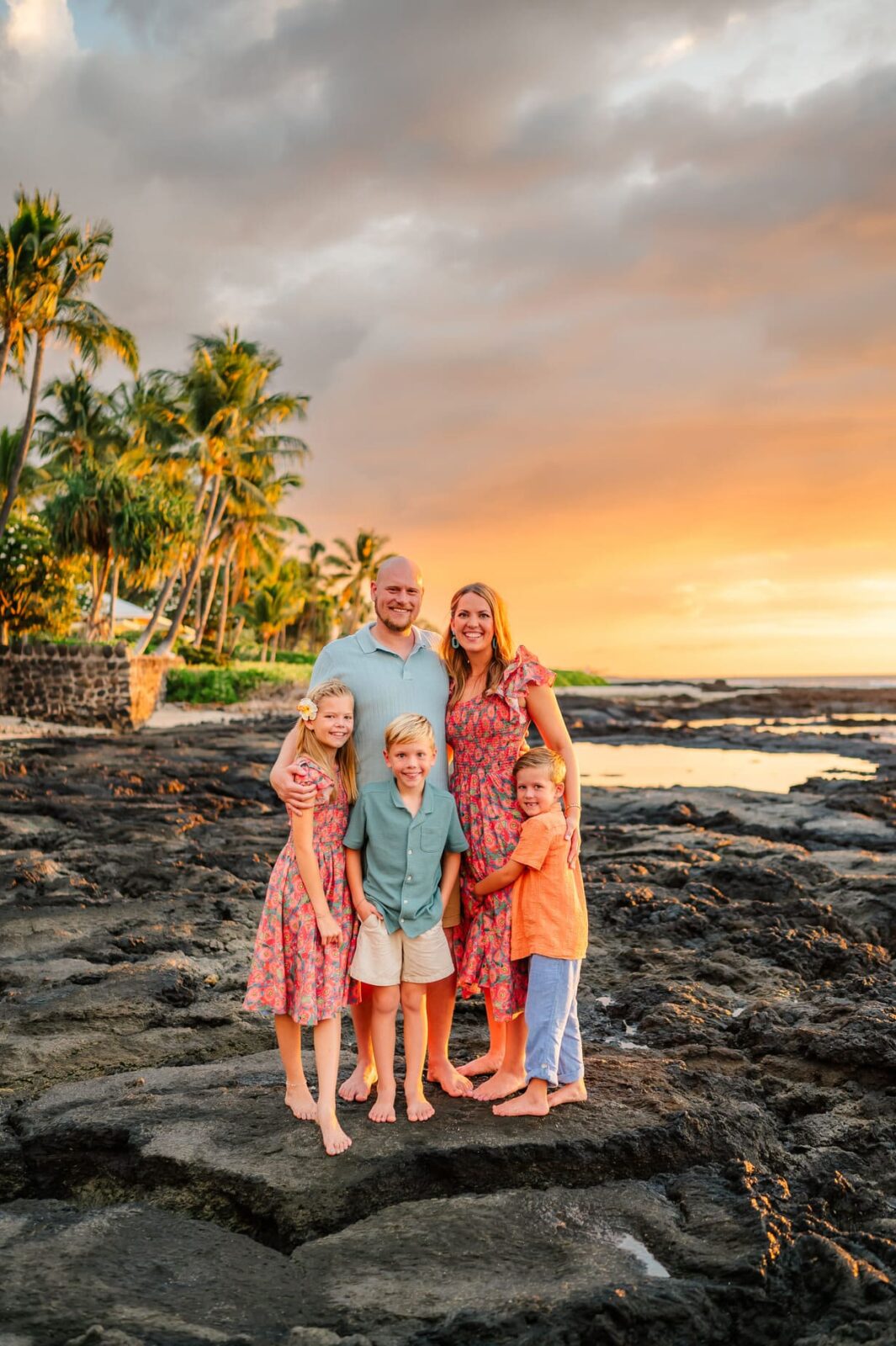 A family of five—two adults and three children—stand barefoot on black rocks near the shore at sunset. They are smiling, dressed in light, summery clothes, with palm trees and the ocean in the background.