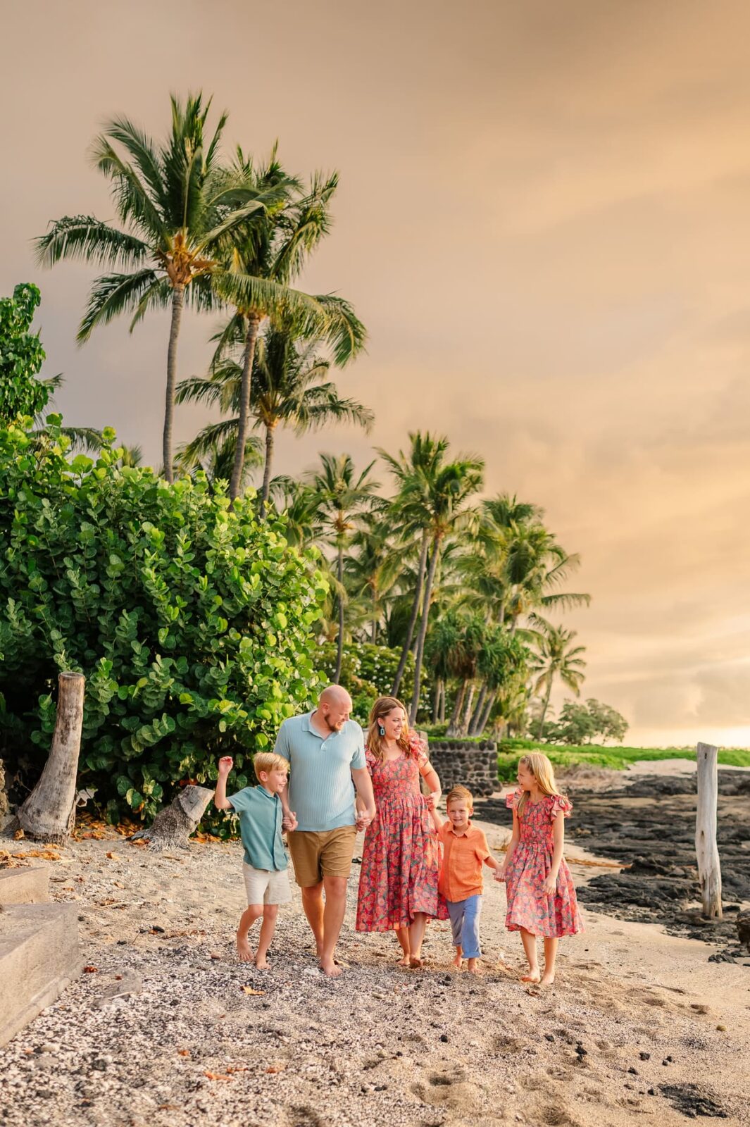 A family of five walks hand-in-hand along a beach lined with palm trees under a golden sunset sky. Two adults and three young children are smiling, dressed in summer clothes, surrounded by lush greenery and sandy shore.