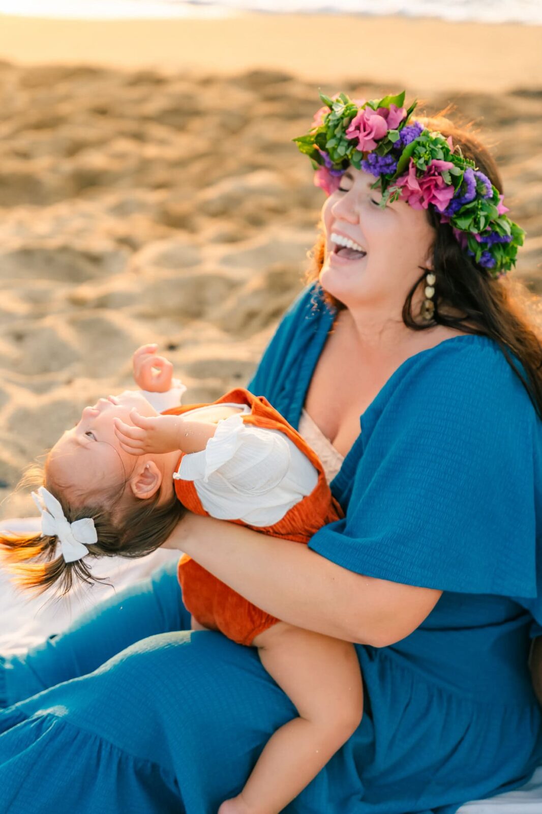 A woman in a blue dress and floral crown laughs while holding a smiling toddler in her lap on a sandy beach. The toddler wears a white shirt with orange overalls and a white bow in her hair.