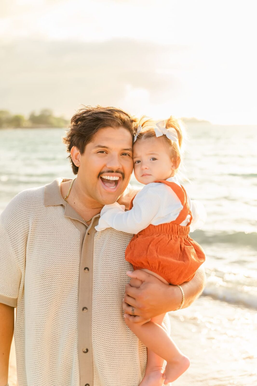 A smiling man with a mustache holds a young girl in his arms on a beach at sunset. The child wears an orange romper and white shirt with a bow in her hair, both looking at the camera with the ocean in the background.