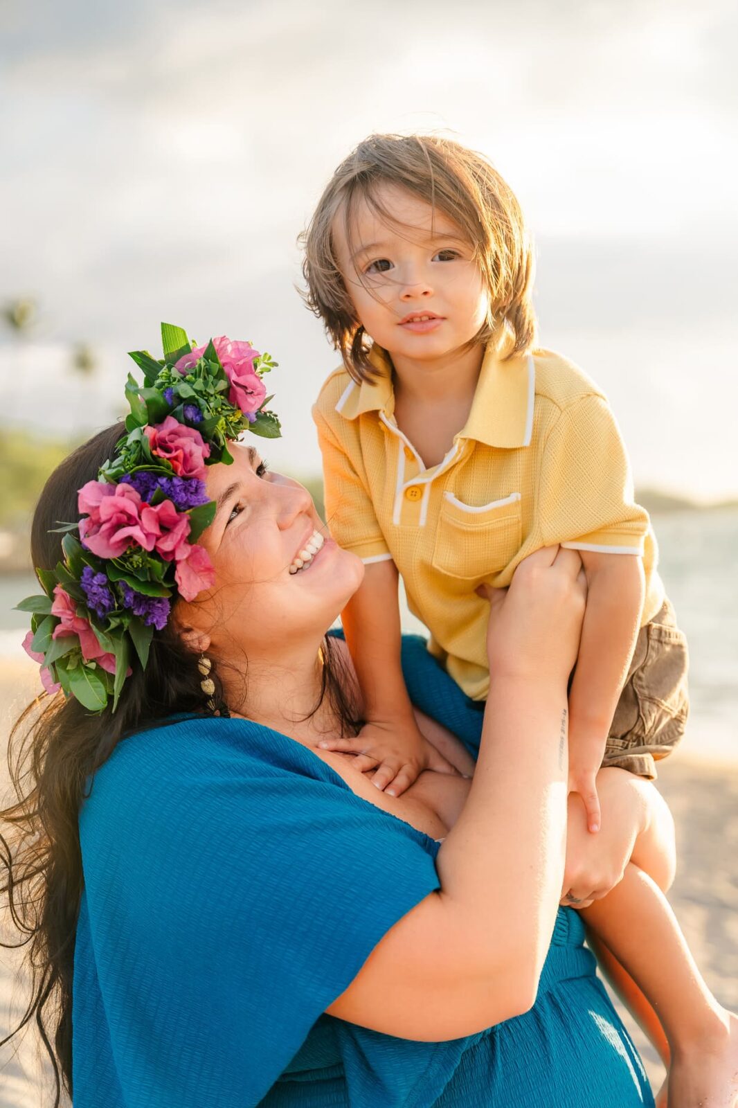 A smiling woman wearing a blue dress and a flower crown lifts a young boy in a yellow shirt at a sunny beach, both looking happy and joyful with the ocean and sky in the background.