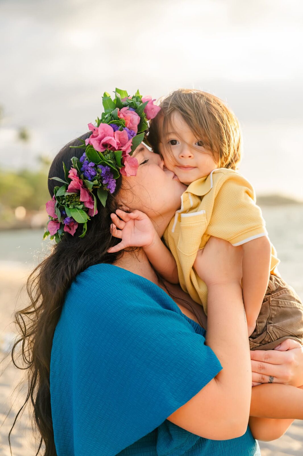 A woman in a blue dress wearing a colorful flower crown kisses a young boy on the cheek while holding him. They are on a beach with soft sunlight and the ocean in the background.