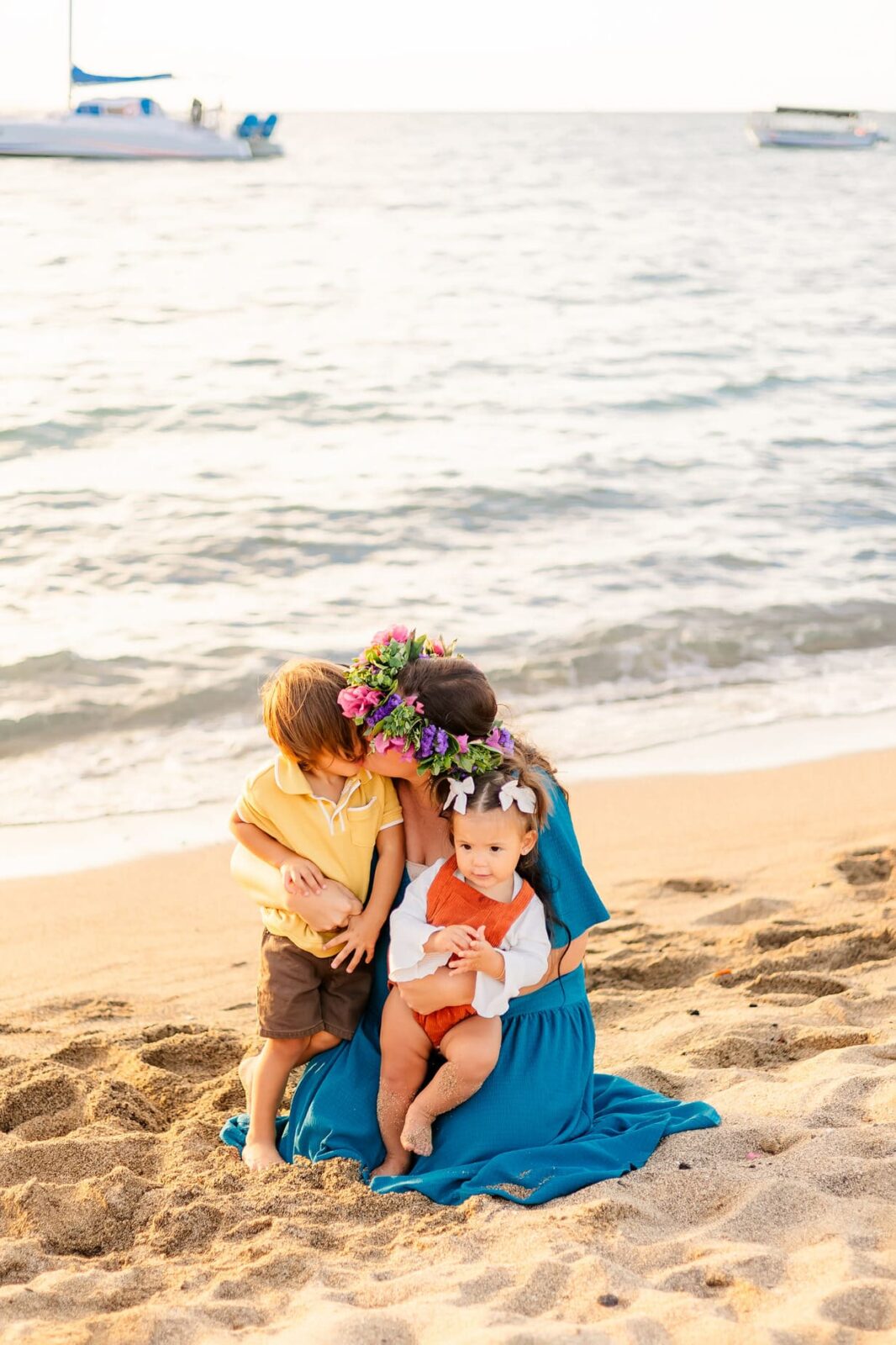 A woman in a blue dress with a flower crown hugs two young children on a sandy beach, with gentle waves and boats visible in the background.