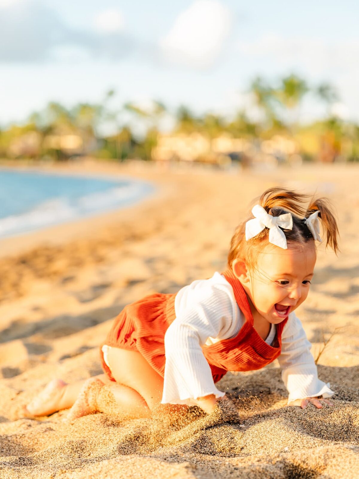 A young child with pigtails and white bows crawls excitedly on sandy beach, wearing a white top and rust-colored overalls, with the sea and palm trees in the blurred background under a sunny sky.